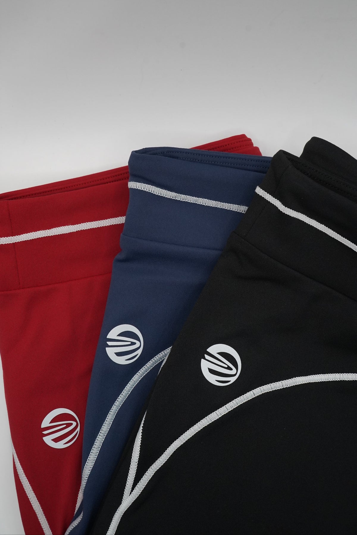 Three pairs of athletic shorts stacked diagonally on a light gray surface, featuring a black pair on top, a navy blue pair in the middle, and a red pair at the bottom. Each pair has a white circular logo on the side with a stylized 'W' design, and white piping borders the edges.
