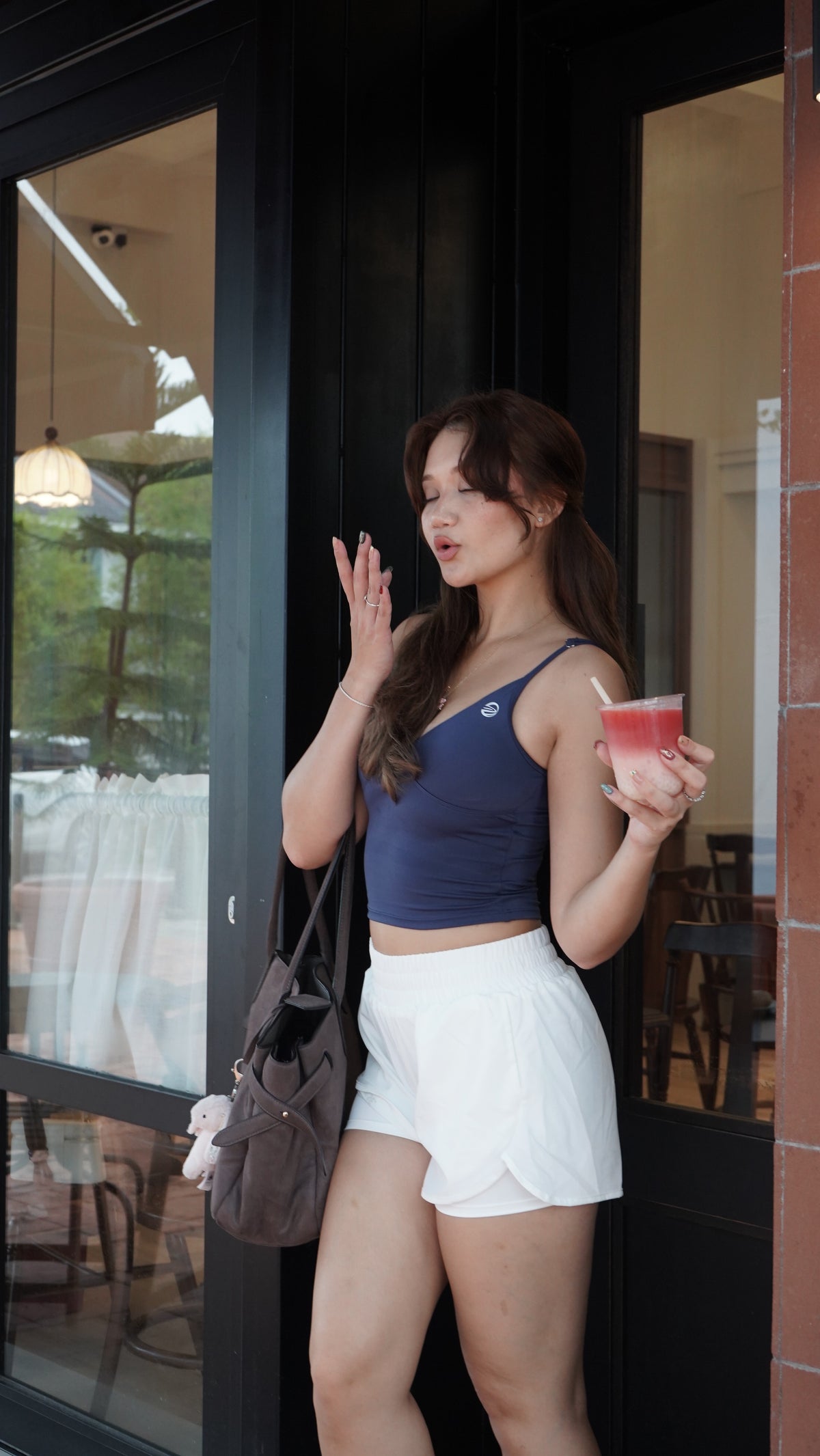 A woman stands outside a storefront wearing a navy blue tank top and white shorts, holding a pink drink and carrying a brown shoulder bag.