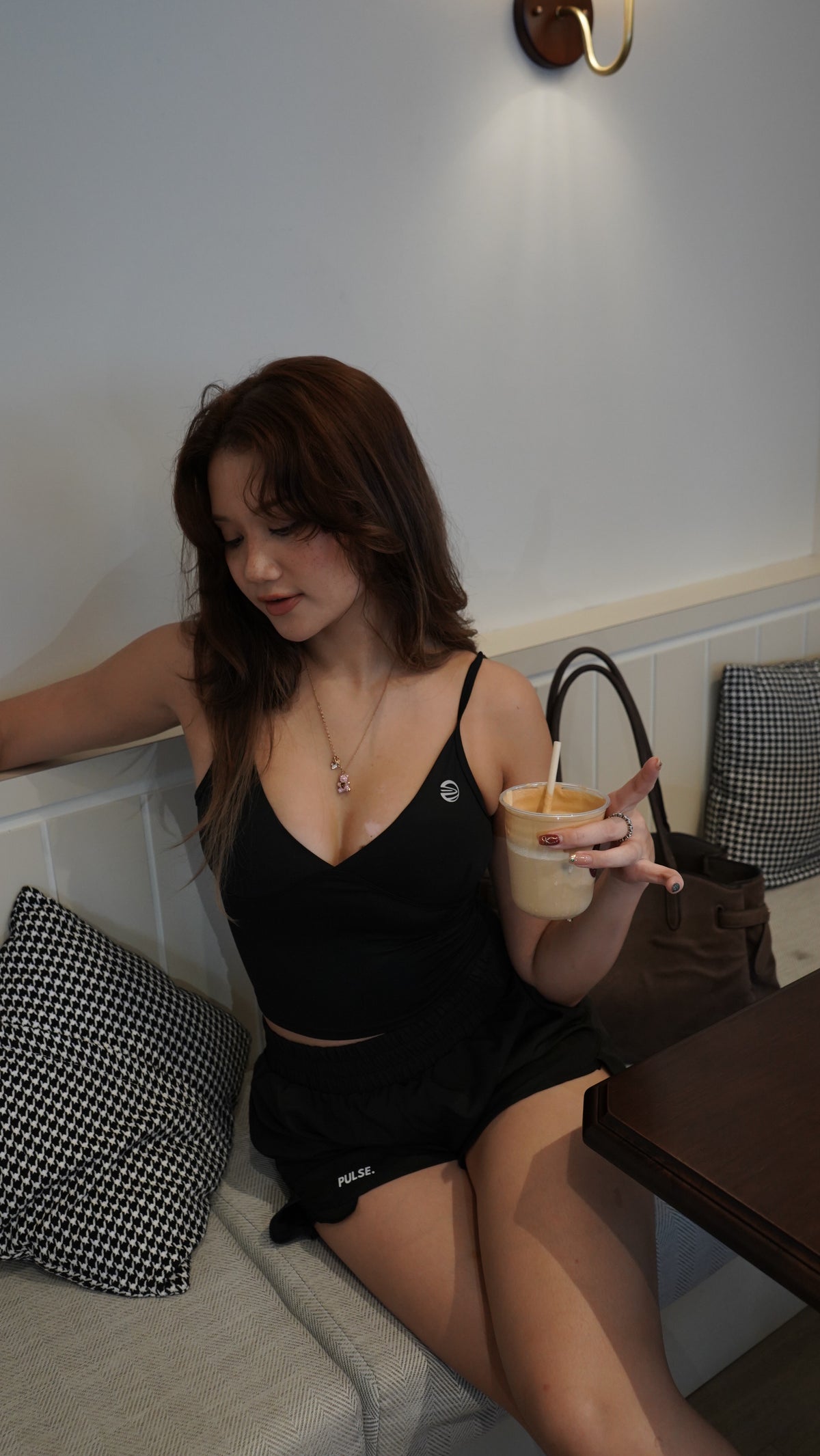 A woman sitting on a light gray sofa in a cafe setting, wearing a black two-piece crop top and shorts set, holding an iced coffee.