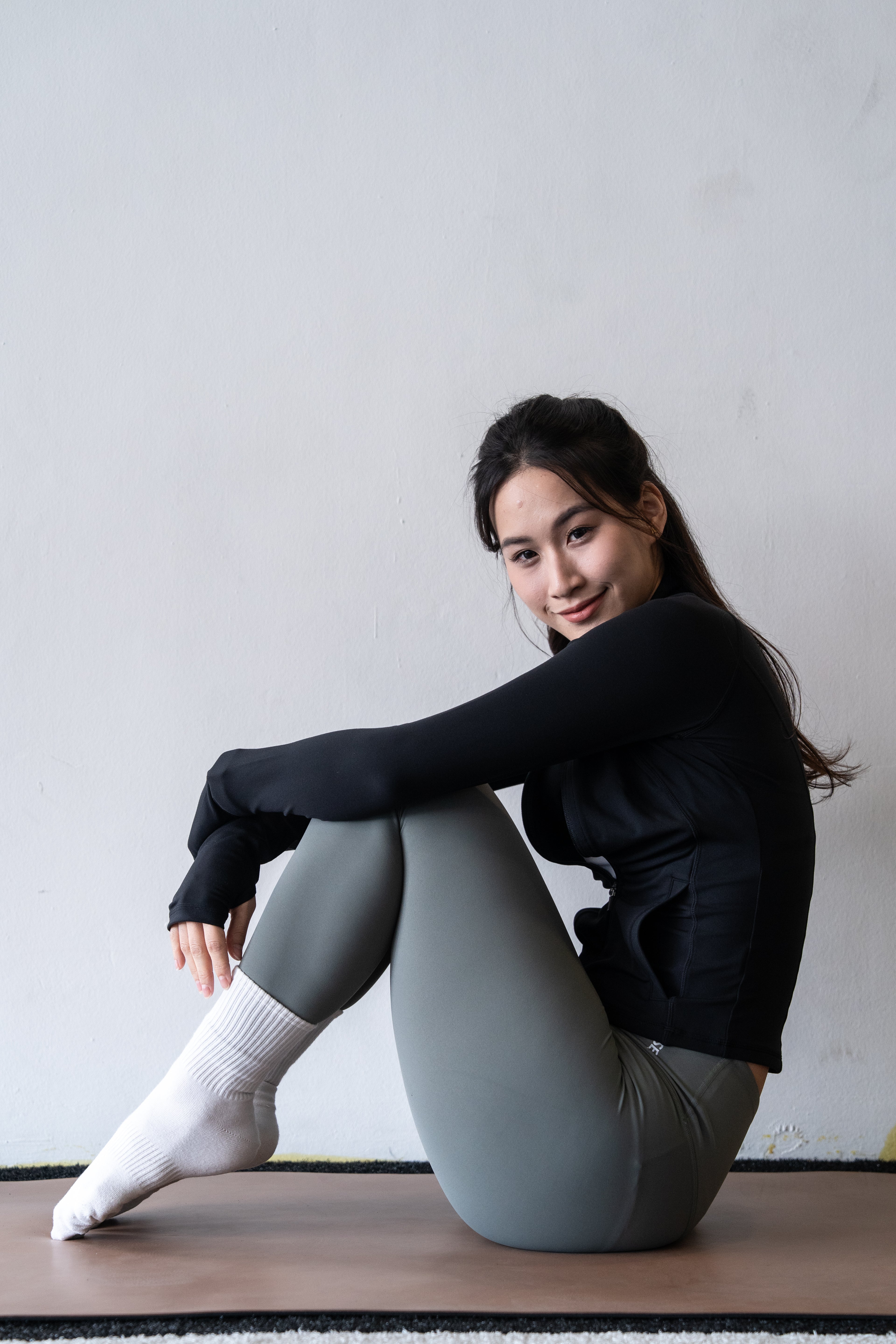 Woman sitting on a wooden floor wearing a black sweater and gray leggings against a plain wall.