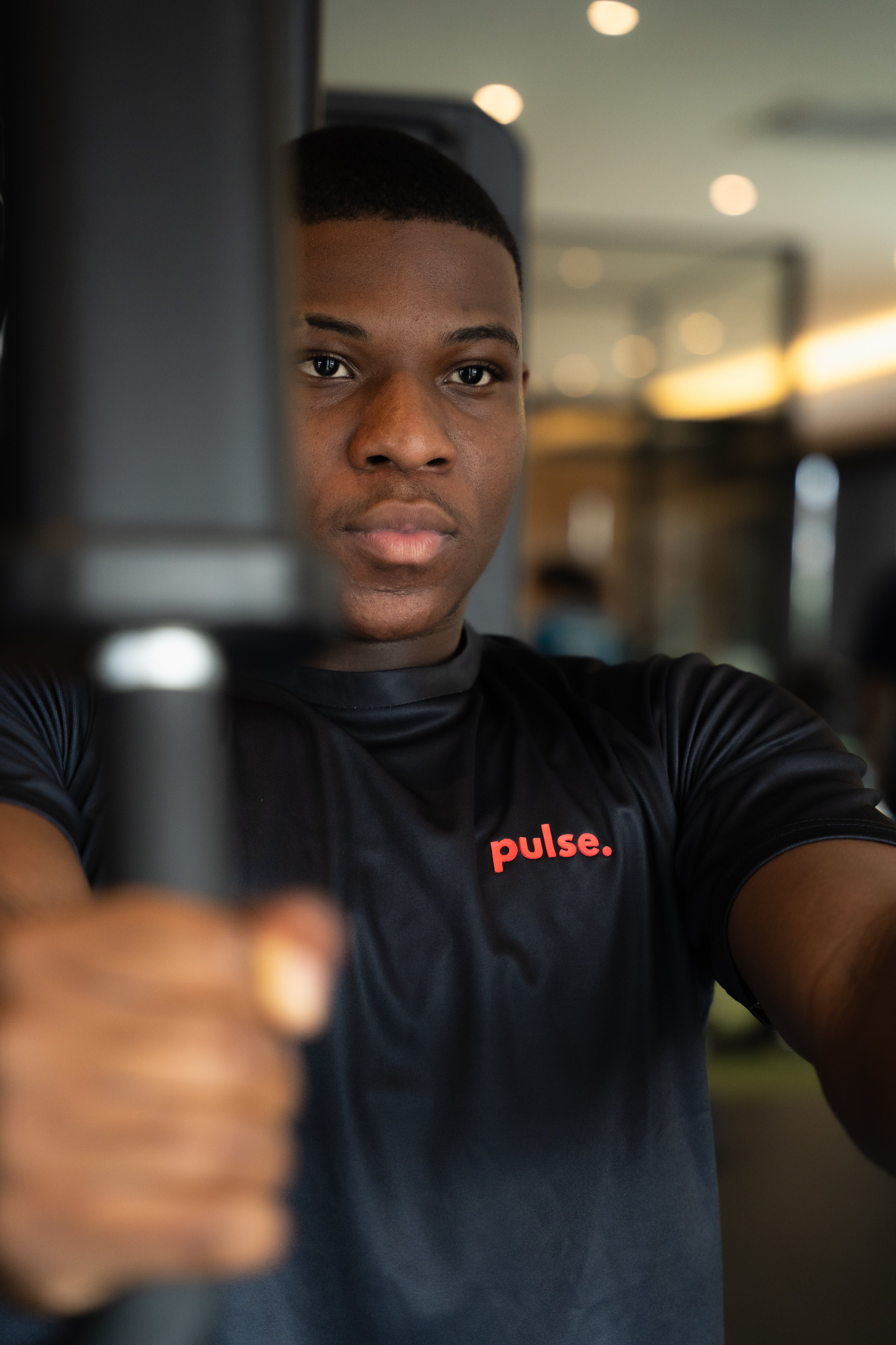 Person wearing a black 'pulse.' shirt holding a large object indoors.